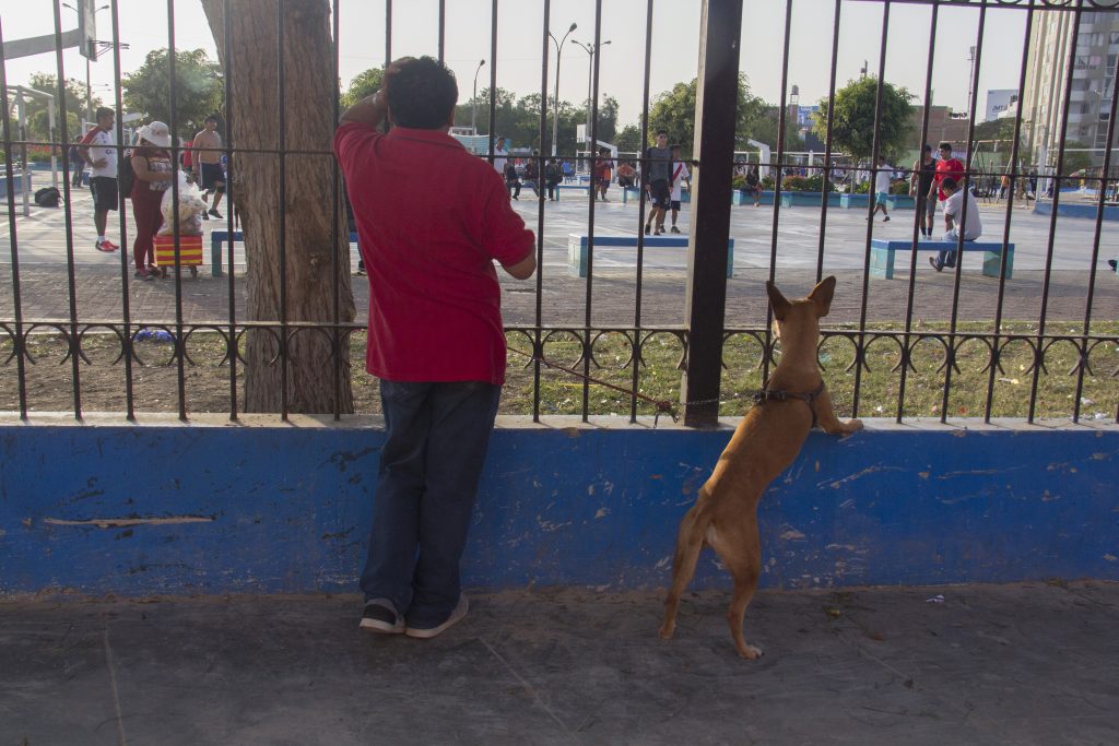Hombre un partido de futbol, junto a un perro, ambos detrás de una reja.
El hombre con camisa roja y pantalón jean color azul, se apoya sobre la reja de metal y el perro posa las dos patas delanteras sobre la base de la reja.