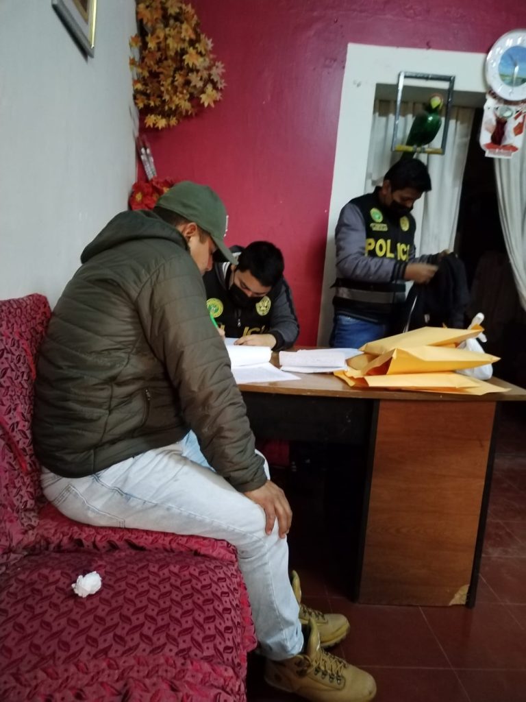 Policía sentado rinde manifestación ante un colega quien anota con un lapicero. Al fondo un agente de policía revisa la mochila del detenido.