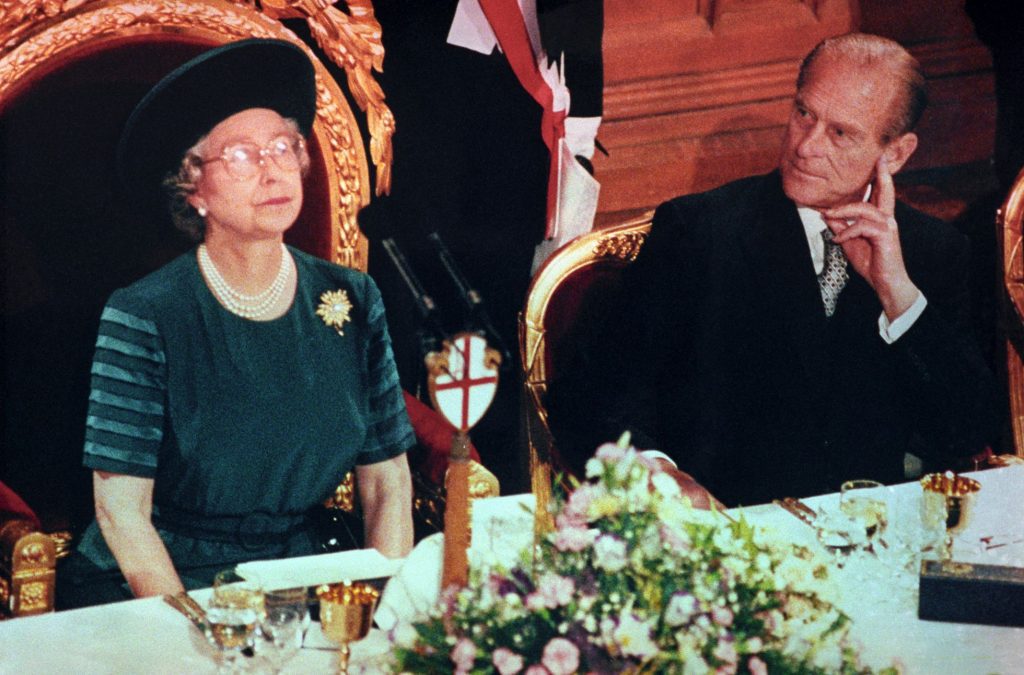 Reina Isabel II sentada con atuendo de color azul, mira al frente, luce collar de perlas de ters vueltas, lentes, pendientes pequeños de color claro. Un prendedor en el pecho del lado izquierdo. Luce sobrero oscuro.
La lado el Rey la observa y éste tiene su mano a un lado de su rostro. Viste con saco oscuro y corbata