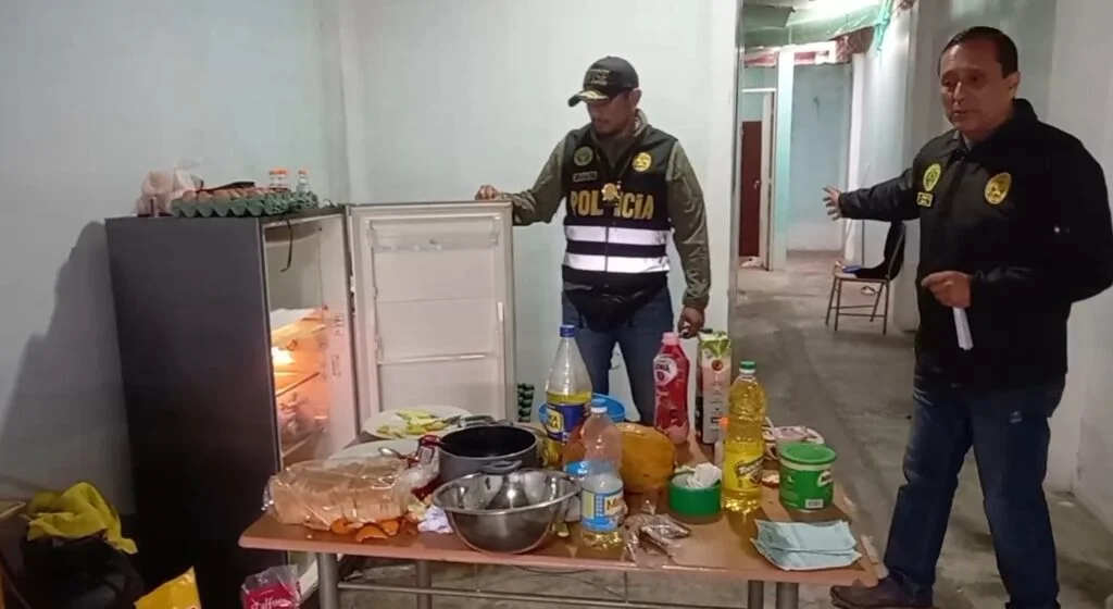 Policía tiene agarrada la puerta de una refrigeradora que está abierta y frente a ella una mesa con alimentos. Otro policía tiene el brazo extendido y muestra el interior de la vivienda