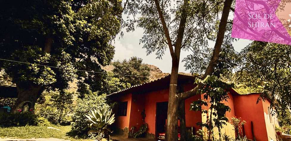 Casa de campo color roja y un árbol a su lado. Al fondo un cerro con árboles.