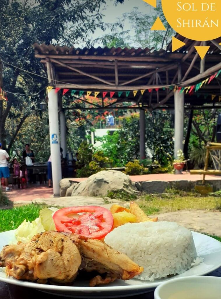 Plato de color blanco con comida compuesta por pollo, plátanos fritos, tomate, arroz y papas fritas. Al fondo dos columnas con un techo