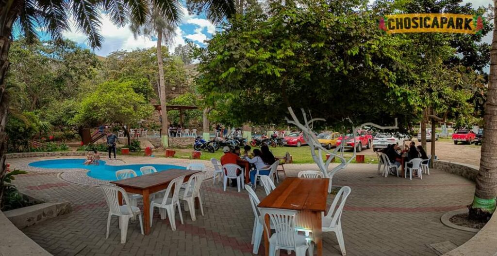 Sillas blancas con un grupo de personas alrededor de una mesa. Al fondo un grupo de autos y un árbol en la escena.
